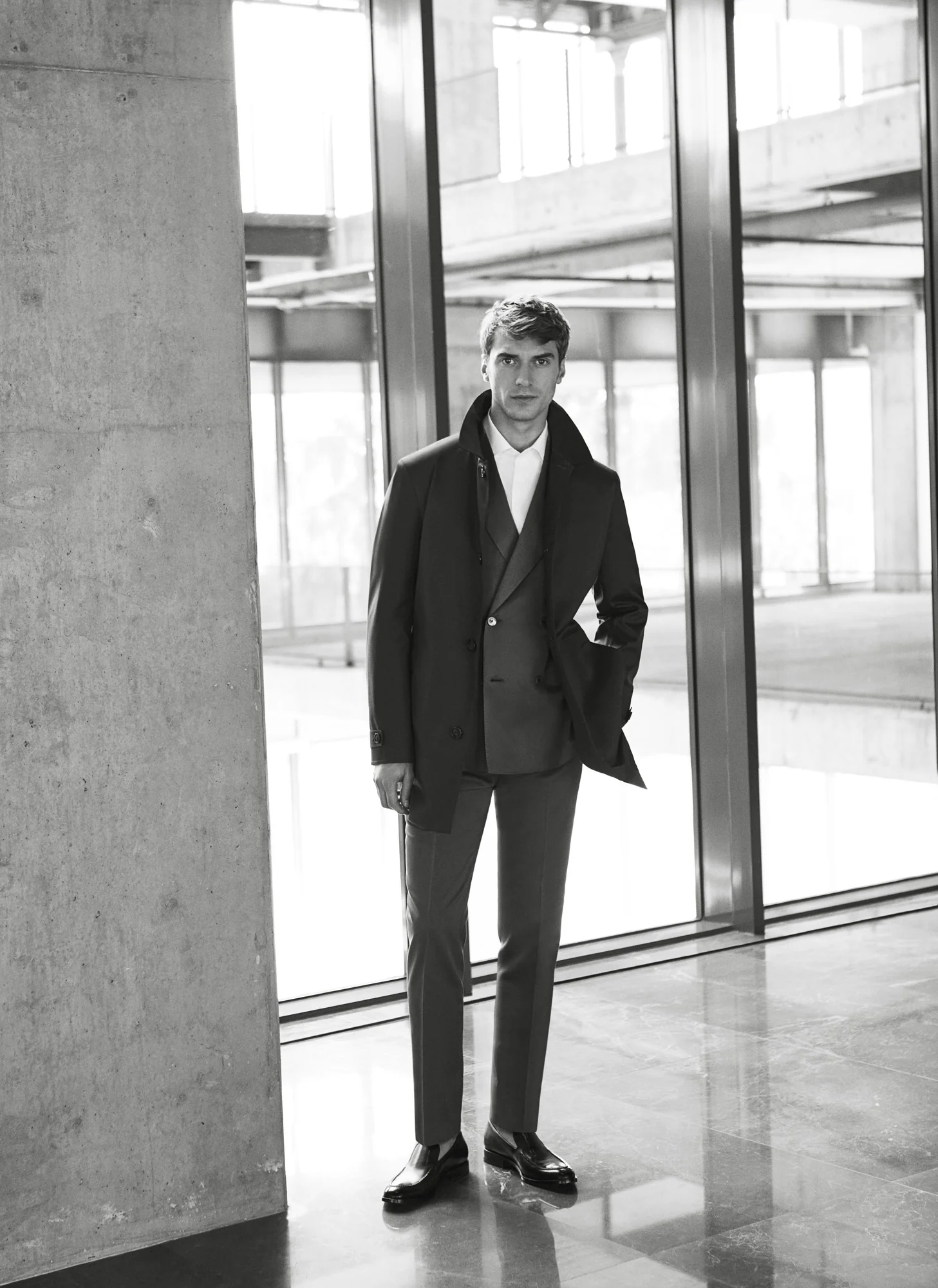 Black-and-white shot of a man in a dark overcoat and suit standing by a concrete pillar and glass wall, for Beymen.
