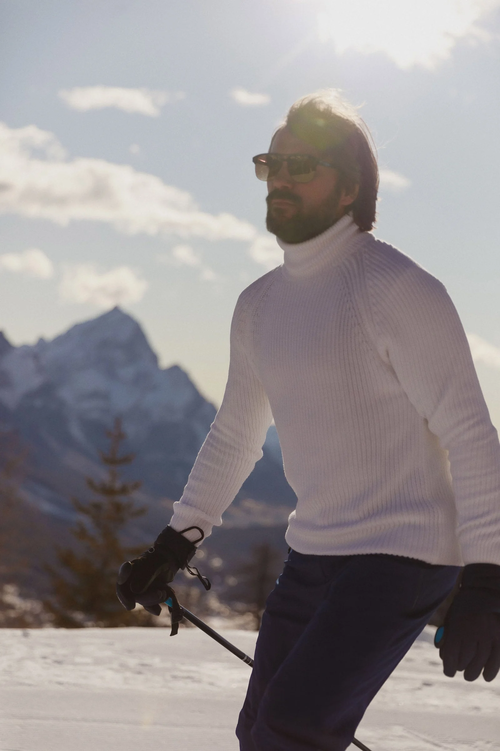 Bearded man in a white knit turtleneck and sunglasses skiing below a snowy mountain peak in winter sun, for Marriott.