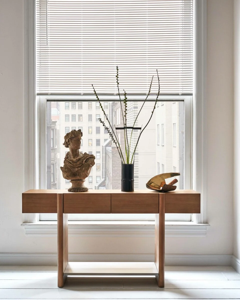 Wooden console table with a classical bust, branch arrangement and brass sculpture before a window overlooking city buildings.