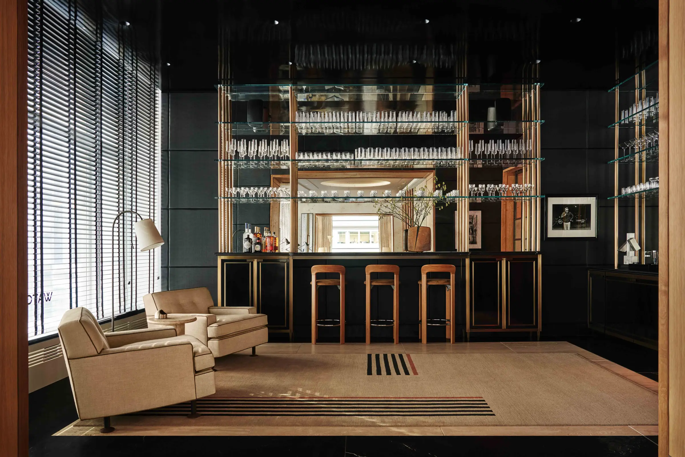 Interior bar lounge with glass shelves of stemware, leather stools and cream armchairs on a striped rug, for WatchBox.