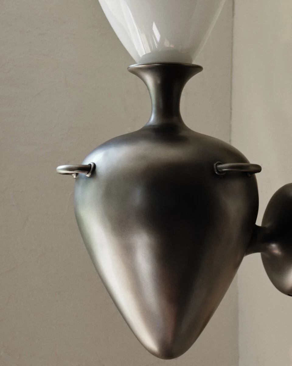 Close-up of a pewter teardrop wall sconce with ring handles and a white glass shade against a plaster wall, for Anna Karlin.