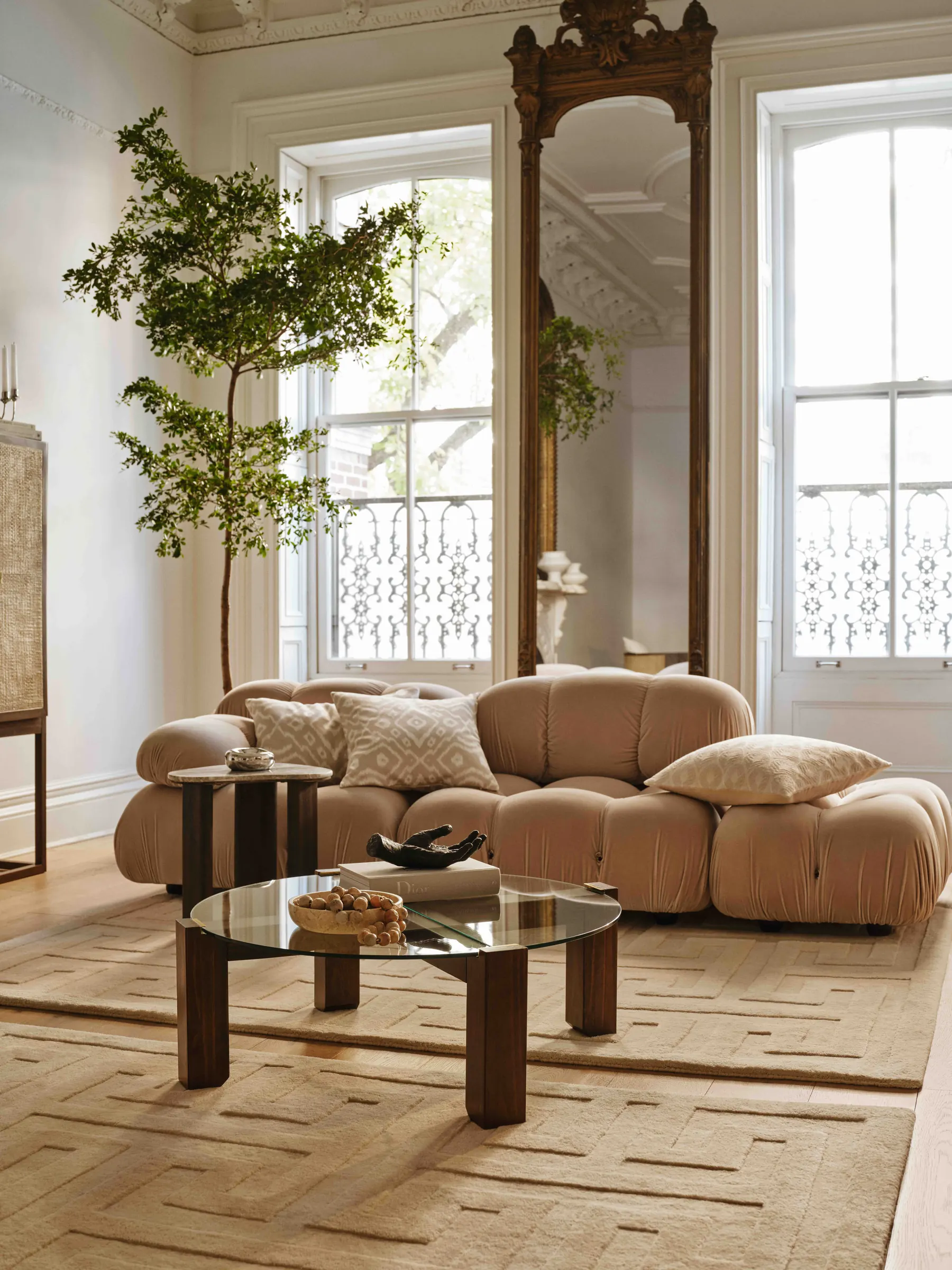 Living room with a beige velvet modular sofa, glass coffee table, gilded mirror and potted tree by tall windows, for H&M Home.