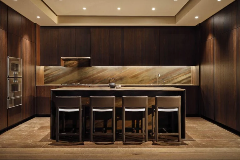 Kitchen with dark wood cabinetry, marble backsplash, island and four leather stools in a residence, for Aman.