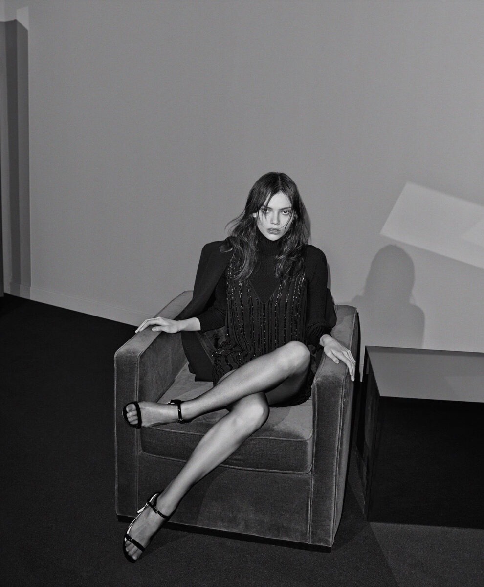 Black-and-white editorial of a model in a beaded black dress and strappy sandals, legs crossed on a velvet armchair.