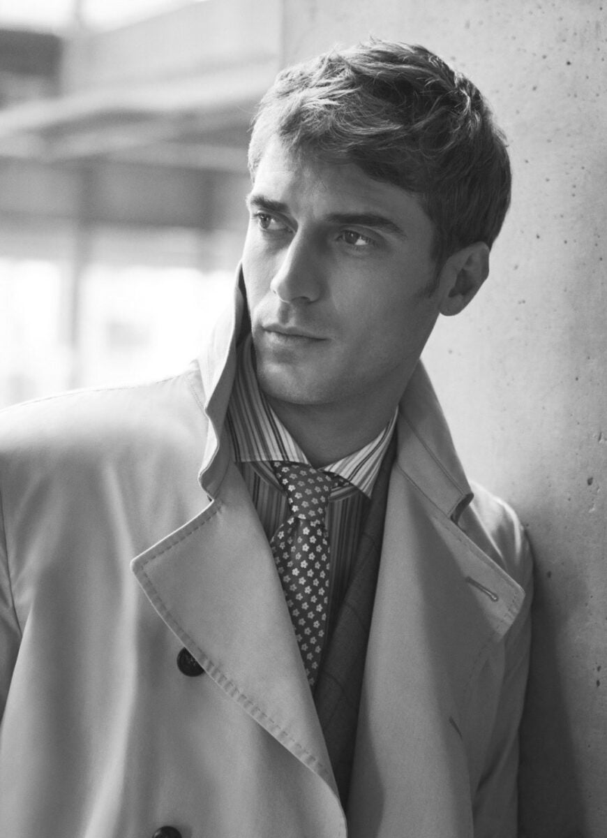 Black-and-white portrait of a man in a trench coat, striped shirt and patterned tie leaning on a concrete wall, for Beymen.