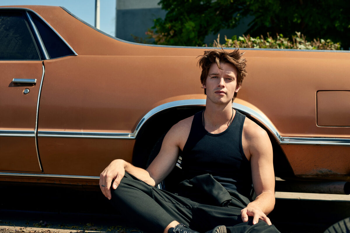 Man in black tank top sitting against a copper vintage car in sunlight, fashion editorial, for Man About Town.