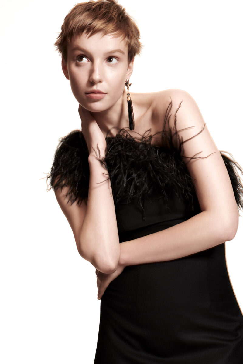 Model with cropped auburn hair in a strapless black dress with feather trim and tassel earrings, on white, for Stella Magazine.