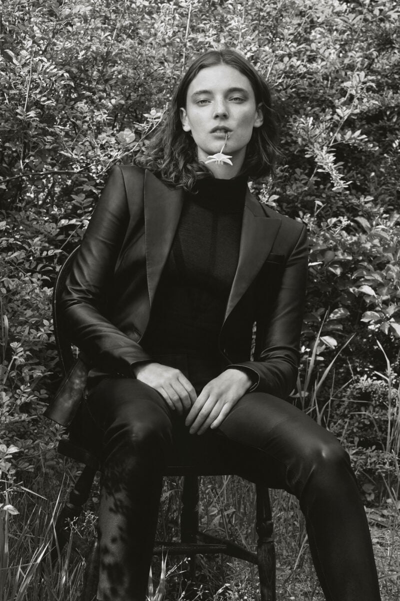 Black-and-white editorial of a model in a leather suit seated on a chair in a garden, a flower in her mouth, for Altuzarra.