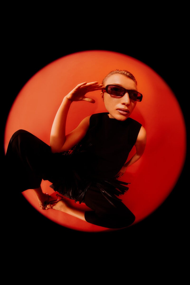 Model in dark sunglasses and black fringed dress crouching in a red circular spotlight, fisheye editorial, for Numéro.