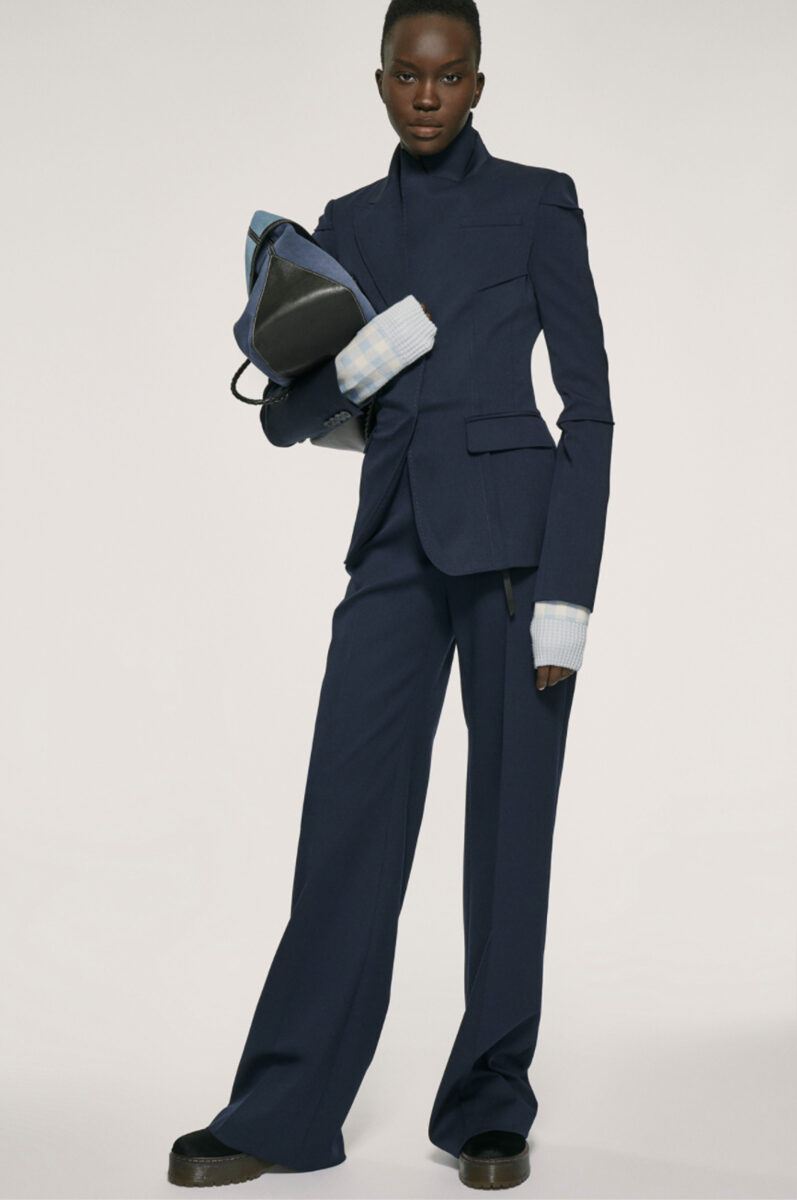 Model in a navy tailored blazer and wide-leg trousers holding a denim and leather bag on a pale backdrop, for Altuzarra.
